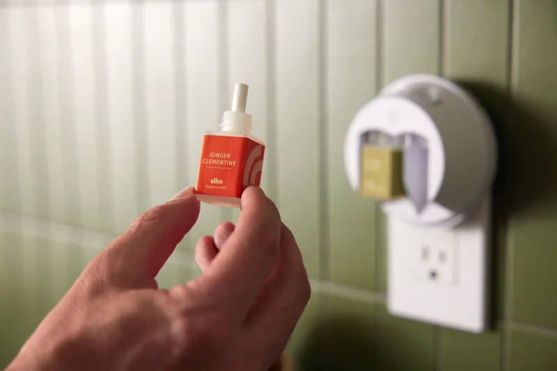 Person holding a ginger clementine fragrance refill next to a plug-in diffuser on a green-tiled wall.