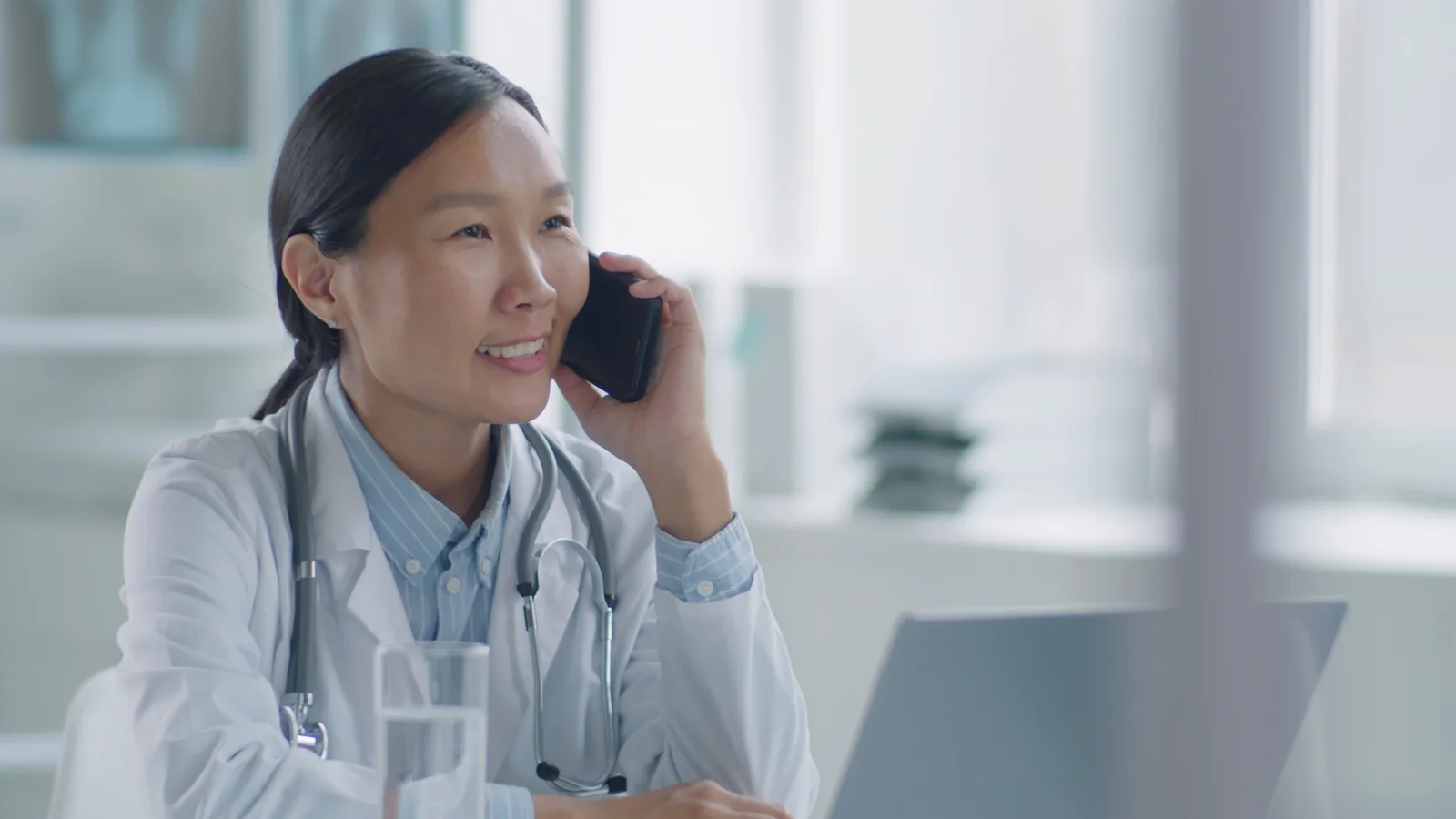 Female doctor wearing a white coat and stethoscope, talking on the phone while using a laptop.
