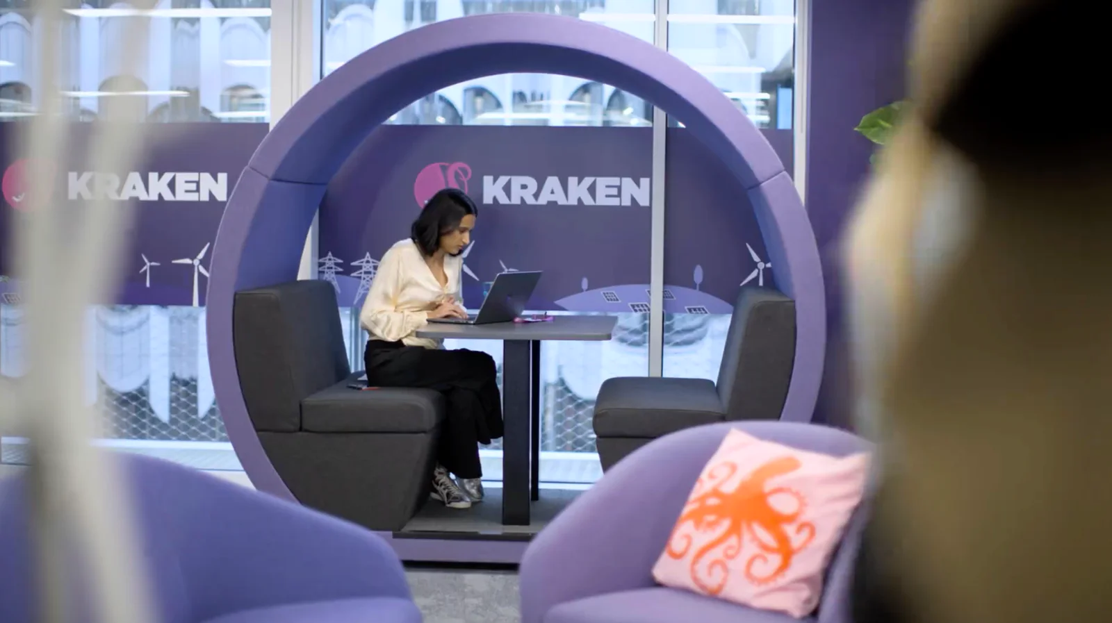 Woman using a laptop in a modern office booth with purple decor and Kraken logo.
