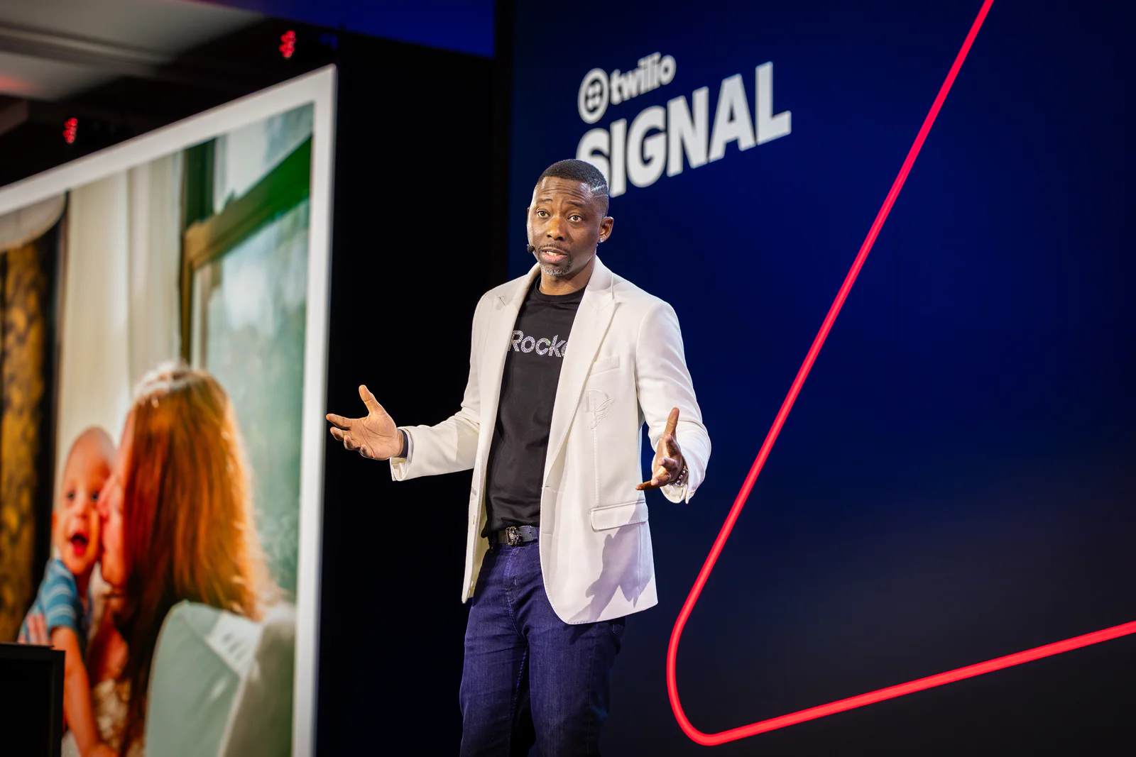 Man wearing a blazer speaks on stage at Twilio Signal conference with an image of a woman and baby in the background.