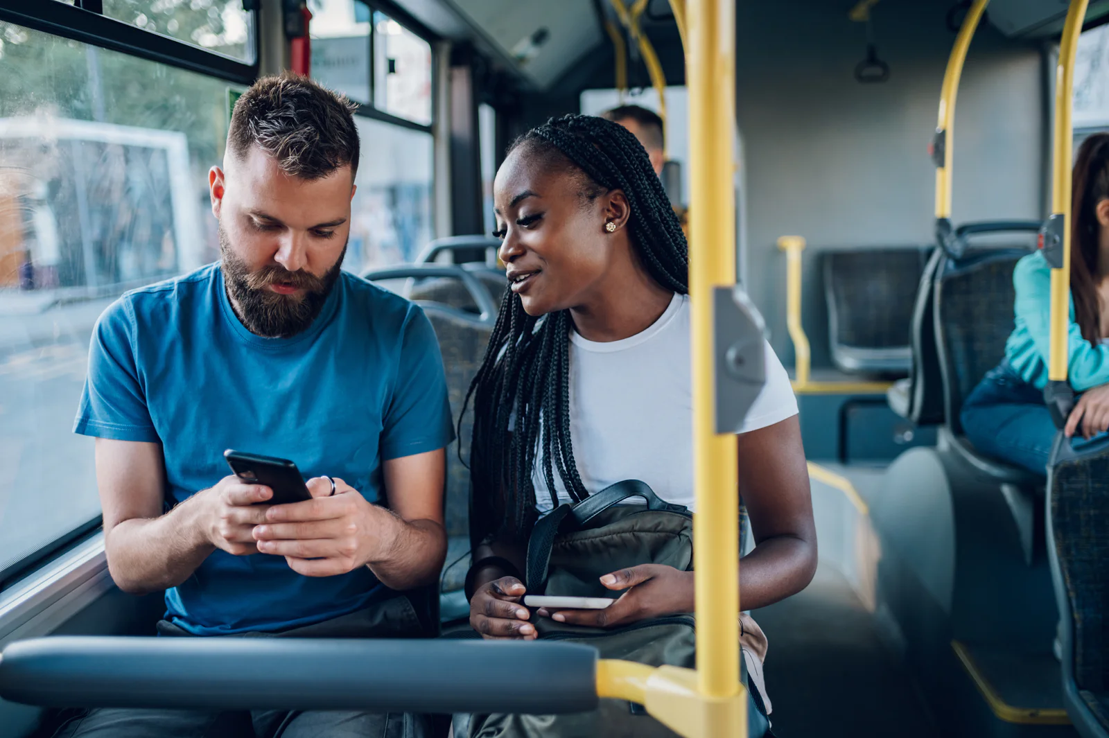 Multiracial friends talking and using a smartphone while riding a bus in the city Smiling diverse friends riding together on a bus and using a smartphone. Two multiracial people enjoying travel or ride on public vehicle and having a conversation. Man typing on a mobile phone.